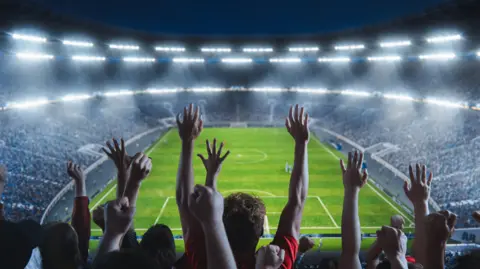 Getty Images Stock image shows a high angle shot of fans cheering for a team in a big stadium during a football match. The team is playing on the pitch which crowds of fans can be seen celebrating, the image shows a wide angle of a sold out arena.