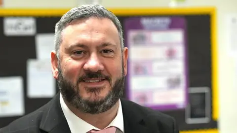 John Fairhall/BBC John Player, who has short grey hair and a dark beard. He is wearing a dark suit with a faint check, a white shirt and pink tie. He smiling and standing in front of a blurred out school notice board