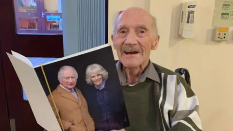 Picture of Dennis Freeman who is 100-years-old. He is sat in a wheelchair and holding a birthday card he received from the King and Queen 