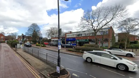 A street view image showing a row of shops in the background and a main road in the foreground with two white cars to the right of the frame.