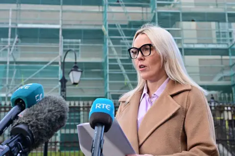 A woman is speaking into a number of news microphones, branded with the logos of various broadcasters. She is reading a statement from a sheet of paper she is holding in both hands. She is wearing a brown jacket, pink shirt and has black glasses and long blonde hair. 