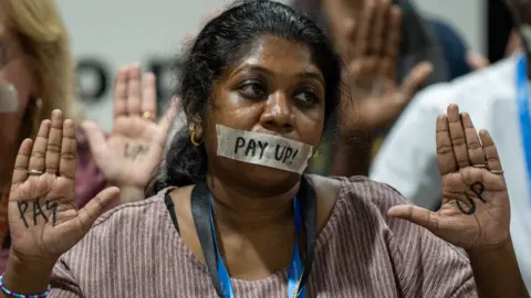 A climate protestor sits with a piece of tape across her mouth with the words 'pay up!' written in black across the tape and on the palms of both hands. 