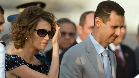 Getty Images Asma al-Assad, who has short curly hair and is wearing sunglasses and a black outfit, touches her hair. Her husband Bashar is wearing a light grey suit and tie. This is a stock photo from 2010.