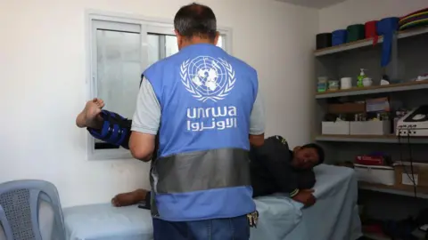 An Unrwa worker in Khan Younis, Gaza. He wears a blue Unrwa vest and is treating a man wearing a leg brace. 