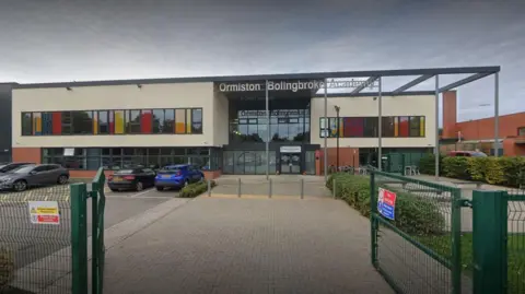 Green gates open onto a path which leads to a large building and car park, a sign on the building reads Ormiston Bollingbroke 