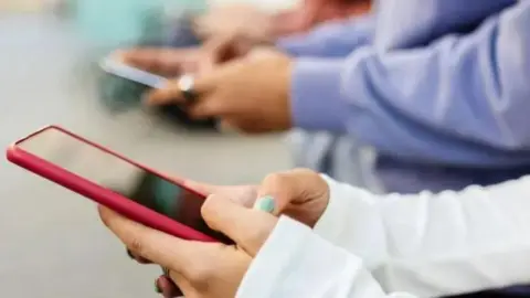 Hands holding a smartphone with a red case. The hands have green nail varnish and the person is wearing a white shirt. Another set of hands are behind holding a phone