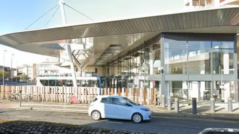 Gloucester Transport Hub from Station Road. Buses can be seen on the forecourt as a white car drives past.