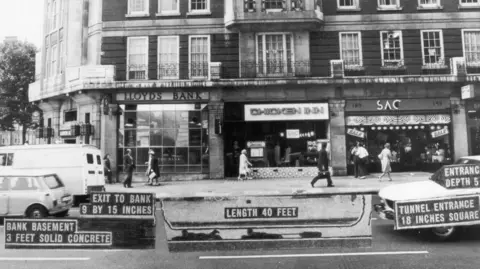 Black and white archive image, showing a row of shops in 1971. Lloyds Bank is on the corner, then a chicken restaurant and then SAC, a leather goods retailer. The image has been notated to show the location of the tunnel beneath the road.