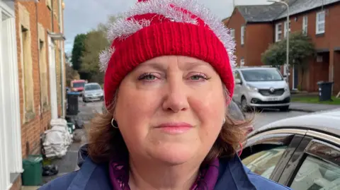 Jane Carr, looking direct to camera and standing on the pavement of a residential street. Jane is wearing a red and silver festive bobble hat and a blue waterproof coat, with a purple coloured top underneath. She has blue eyes and her light brown hair can be seen emerging from under the red hat. Immediately to the right of her in the image is the side of a car. A silver van is parked further away behind her on the opposite side of the road. To the left of the image are the frontages of terraced houses. On the pavement are some small foodwaste bins, as well as a pile of sandbags.