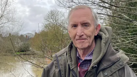 Ben Schofield/BBC David Adams looking direct to camera and standing in front of a body of water and several trees, most of which have shed their leaves. The body of water is the River Ouzel, which is at flood. Richard has grey hair and is wearing a purple checked shirt, dark red v-neck sweater, khaki hooded top and a heavy, khaki waterproof overcoat. 