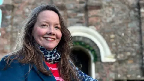 Caroline Devlin, a woman with brown hair wearing a red jumper, blue blazer and scarf in front of the entrance of the church, smiling.