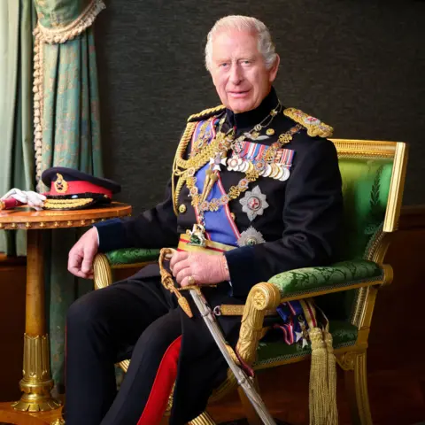 PA The photograph of King Charles, in which he is seated in ornate green and gold chair, wearing his full dress uniform with a ceremonial sword and ceremonial hat sitting on a table next to him