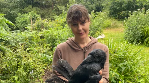 A woman in a brown hoodie holds a dead raven in her hands as she looks at the camera, in a field with lots of trees and bushes