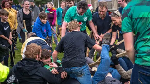 A number of players on the ground on top of each other while other players hold out sticks trying to get the ball. Other watch on smiling.