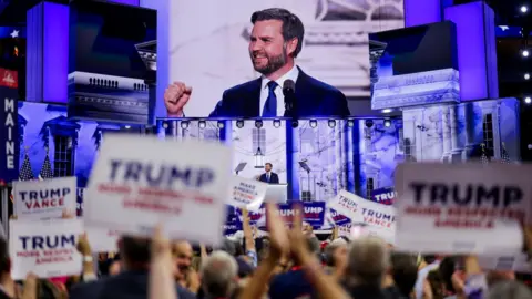 JD Vance seen on a big screen as supporters carrying signs reading TRUMP