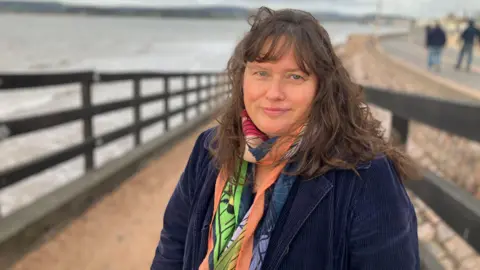 BBC Nicky Nicholls stands on a slipway by Exmouth beach. She has a multi-coloured scarf on and a navy blue corduroy jacket on. She has long brown hair. The tide is in in the background. 