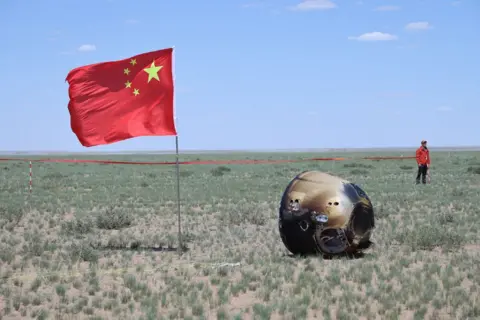 Xinhua/REX/Shutterstock The retrieval site of the capsule of the Chang'e-6 probe in Siziwang Banner, north China's Inner Mongolia. China's flag is on the left of the probe capsule which appears on the ground on its side, with heat marks on the exterior. A man in red stand near a cordon tape in the background