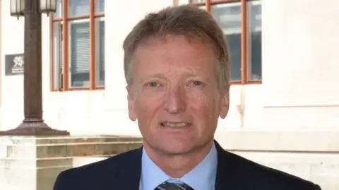 Sir Dr Frank Atherton wears a suit and smiles at the camera. He is standing in front of a white stone government building