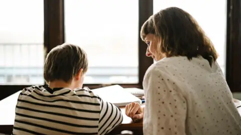 A boy and a woman are sat at a table with their backs to us. The boy is wearing a black and white striped top and looking at a notebook. The woman is wearing a white top and looking down at the boy's notebook.