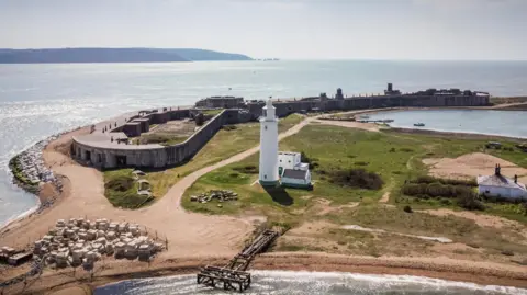 Hurst castle, with its low ceilings and perimeter spanning the tip of the shingle bank, and a white lighthouse, pictured at the tip of Hurst Spit in the foreground against the sea in the background. 