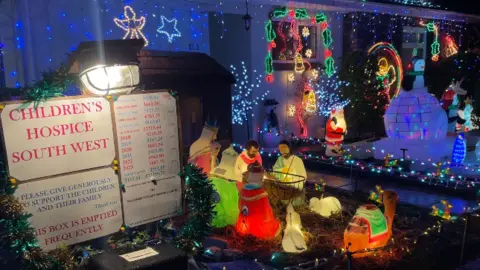 A sign has been placed outside of the home. The sign, which is illuminated, states 'Children's Hospice South West'. Please give generously to support the children and their family, thank you. This box is emptied frequently.'
A number of Christmas decorations can be seen next to the sign including a large nativity scene.