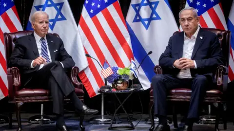 In front of US and Israeli flags, Joe Biden sits with his legs crossed while to his left sits Israeli Benjamin Netanyahu who clutches his hands in front of his chest