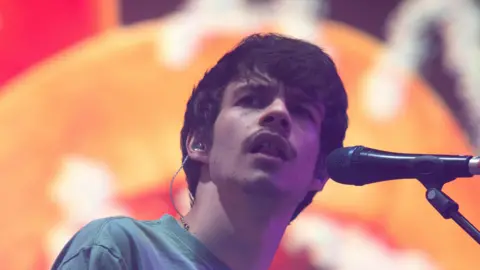 Rex Orange County has medium length brown hair and brown stubble. He is singing into a mic, with an orange background out of focus behind him.