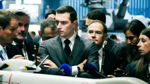  DANIEL DORKO/Hans Lucas/AFP Jordan Bardella (C), president of the National Rally party looks at an Airbus military drone model at the Eurosatory international exhibition for the land and air land defence and security industry in Villepinte