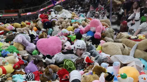 Ice hockey player lying in pile of stuffed toys on ice rink