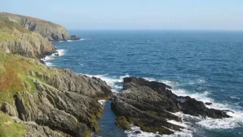 CROGGA A coastline of rocky cliffs with green grass atop them, and blue sea stretching off into horizon on the right side of the image. 
There is a large rocky outcrop at the bottom of the cliffs. 