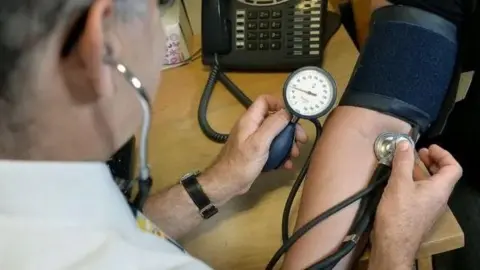 Man holding a blood pressure stethoscope to another person's inner arm while looking at the dial of the unit