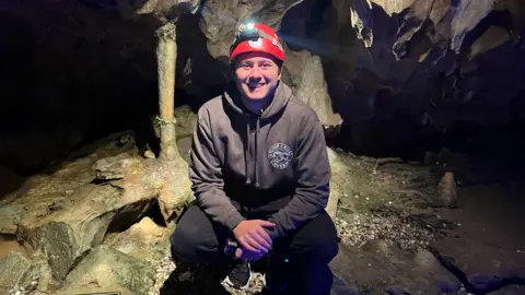 A man, who wears a red helmet and grey hoodie, crouches down and smiles inside a cave.