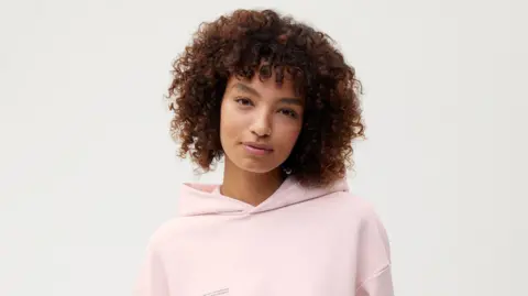 A model with short curly hair wearing a pale pink hoodie.