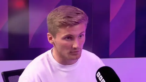 Taylor Moore wears a white t-shirt and sits in front of a black microphone with the BBC logo on it.