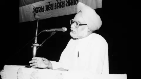 A back-and-white image of former Indian prime minister Manmohan Singh speaking in front of a microphone