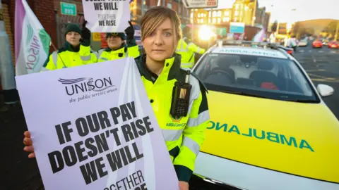 PA Media An ambulance worker in florescent jacket holds a sign saying "if our pay doesn't rise we will" standing beside an ambulance estate car. Behind her are other striking ambulance workers. 