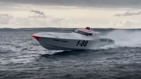 A high-speed powerboat cuts through the water, labelled with the number "1-88" and the name "Double Trouble" on its side. The aerodynamic vessel has water spray behind it, suggesting it is moving at a high velocity. The boat has a white body with a bright red tip on the bow, and sponsors' logos are visible along its side. The sea appears slightly choppy under a cloudy sky, with land visible in the background.