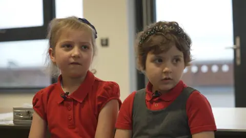 Betsy and Lacey sitting beside eachother. Lacey wears a red top and Betsy wears a red top and a grey pinafore.