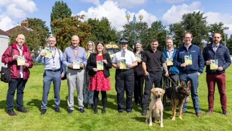 Merseyside Police Taking The Lead campaign launch - Councillors and police represntatives and dog owners holding two dogs gather on grass and hold up campaign leaflets 