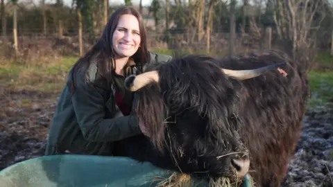 Chloe Lucas, crouching down next to a Highland cattle with dark coloured fur. Chloe is wearing a dark green jacket and is smiling at the camera.