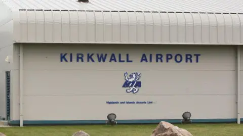 A general view of Kirkwall Airport. The terminal building is grey, with the name and a lion motif in blue and white on the side. There is a patch of green grass in front.