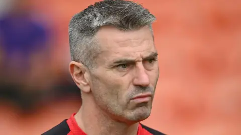 A man looking off camera, only his head and the tops of his shouldres are visible. He has short greying dark hair and stubble. He is wearing a black and red top. The background is blurred but it appears to be the stand of a sports ground