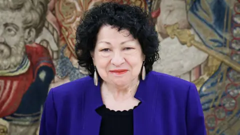 Sonia Sotomayor, who has a short curly bob of black hair, and is wearing a deep blue jacket over a black blouse, with hanging feather-like earrings, poses for a photo during a visit to Spain