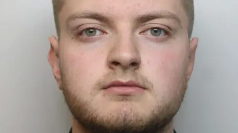 Bearded Mason Hodgson, wearing a black shirt, looks unsmiling and straight ahead in his police mugshot