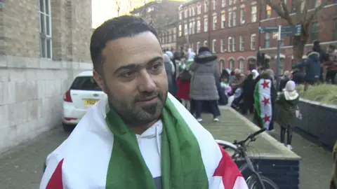 Man draped in Syrian flag outside broadcasting house, red brick buildings in background, with crowd celebrating in background