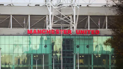 EPA A woman in a blue rain coat with a black rucksack crosses a road, with the Old Trafford stadium seen towering behind her. 