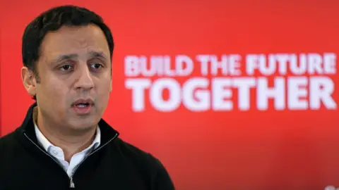 PA Media A man with dark hair, wearing a black jumper over a white shirt, speaks in front of a red Labour background in a medium close-up shot 