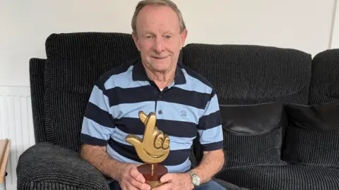 A man in his eighties sits on a black sofa in a living room. He is holding a brass-coloured trophy in the shape of the National Lottery logo