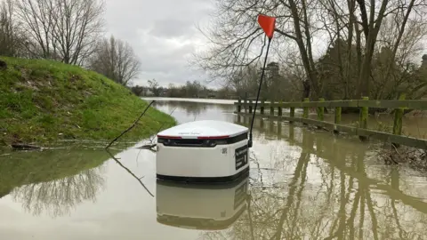 DJ McLaren/BBC A starship delivery robot submerged in flood water. The robots, which are like smart cool-boxes on wheels, are used to autonomously deliver groceries in the neighbourhood. This robot's wheels are all submerged beneath muddy water. It is mostly white, with two black bands running around its top and bottom. The words "open here" can just be made out on its roof or lid. It also has a flag pole coming up from its rear, right hand corner. At the top of the pole is a triangular orange flag. Elsewhere in the picture is a small grassy bank, a wooden fence running from the right of the image into the middle distance, and some bare trees. In the far distance some flooded playing fields can be made out.