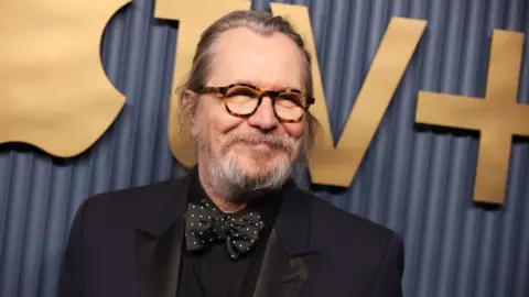 Gary Oldman in a black and blue suit with a black and white polka dot bow tie standing in front of a navy-coloured wall with Apple TV+ logo in gold